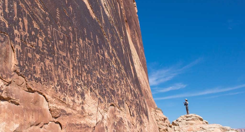 A person stands looking at a red wall containing petroglyphs 